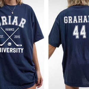 May include: Navy blue t-shirt with white text and graphics. The front features "Briar Hawks University" with crossed hockey sticks and a puck. The back displays "Graham 44". A casual, oversized tee.