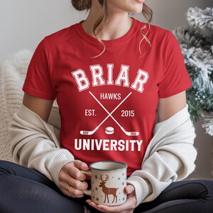 May include: A red t-shirt with a white graphic featuring the text "Briar Hawks University" and crossed hockey sticks. The shirt includes the year "2015" and is being worn by a person holding a mug with a deer design.