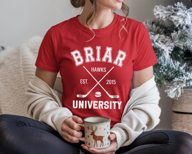 May include: A red t-shirt with a white graphic featuring the text "Briar Hawks University" and crossed hockey sticks. The shirt includes the year "2015" and is being worn by a person holding a mug with a deer design.
