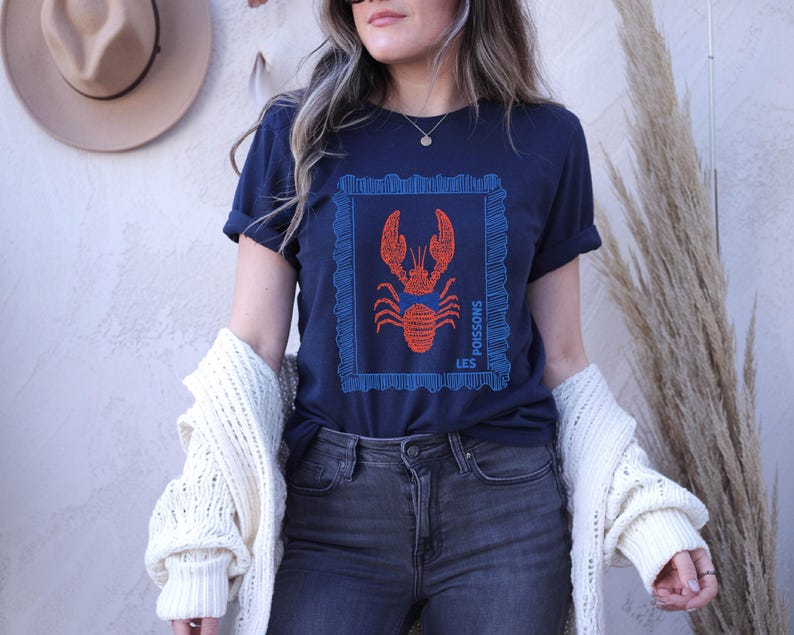 May include: Navy blue t-shirt featuring a red lobster design within a blue frame. The words "LES POISSONS" are printed vertically on the left side of the frame. The shirt is a casual style.