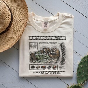 May include: Cream-colored t-shirt featuring a detailed illustration of a sea otter with the text "Sea Otter Enhydra Lutris." The graphic includes facts, primary food sources, and the Monterey Bay Aquarium logo. A straw hat is in the upper left corner.