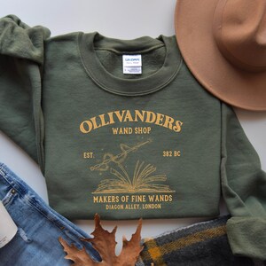 May include: Green sweatshirt with a gold graphic of Ollivanders Wand Shop, featuring a book with a wand and stars. The text reads "Ollivanders Wand Shop, Est. 382 BC, Makers of Fine Wands, Diagon Alley, London."