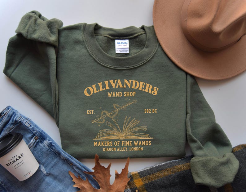 May include: Green sweatshirt with a gold graphic of Ollivanders Wand Shop, featuring a book with a wand and stars. The text reads "Ollivanders Wand Shop, Est. 382 BC, Makers of Fine Wands, Diagon Alley, London."