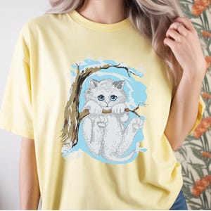 Könnte beinhalten: Hellgelbes T-Shirt mit einem weißen Kätzchen, das an einem Ast hängt. Die Katze hat blaue Augen und ist vor einem hellblauen Hintergrund platziert. Das T-Shirt hat einen lässigen Stil.