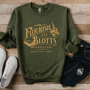 May include: A green sweatshirt with a graphic print of the text "Flourish and Blotts Booksellers Diagon Alley, London" in a vintage style font. The graphic also includes a quill pen and stacked books.