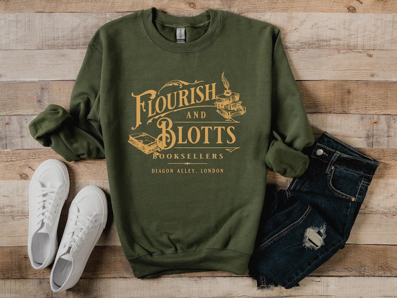 May include: A green sweatshirt with a graphic print of the text "Flourish and Blotts Booksellers Diagon Alley, London" in a vintage style font. The graphic also includes a quill pen and stacked books.
