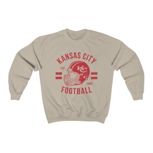 Pode incluir: Uma camisola bege com um logotipo vermelho de estilo vintage para os Kansas City Chiefs. O logotipo apresenta um capacete de futebol com as iniciais da equipa "KC" e o texto "Kansas City Football" com o ano de funda&ccedil;&atilde;o da equipa "1960" e "Est." para estabelecido.