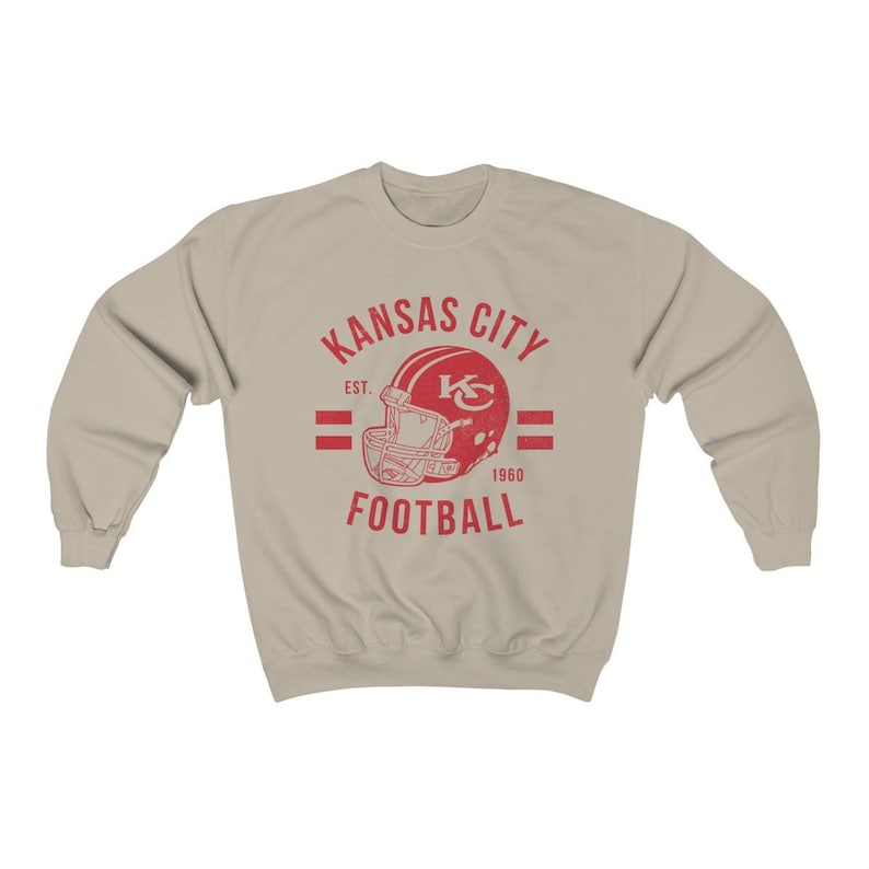 Pode incluir: Uma camisola bege com um logotipo vermelho de estilo vintage para os Kansas City Chiefs. O logotipo apresenta um capacete de futebol com as iniciais da equipa "KC" e o texto "Kansas City Football" com o ano de funda&ccedil;&atilde;o da equipa "1960" e "Est." para estabelecido.
