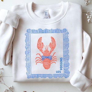 May include: White sweatshirt with a screen-printed design of a red lobster wearing a blue bow tie, inside a blue rectangular frame. The words "LES POISSONS" are printed in blue.