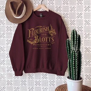 May include: Burgundy sweatshirt with a gold graphic of the fictional bookstore "Flourish and Blotts" from the Harry Potter series. The graphic includes the text "Flourish and Blotts Booksellers Diagon Alley, London".