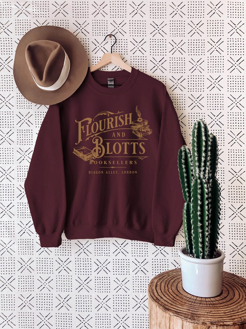 May include: Burgundy sweatshirt with a gold graphic of the fictional bookstore "Flourish and Blotts" from the Harry Potter series. The graphic includes the text "Flourish and Blotts Booksellers Diagon Alley, London".