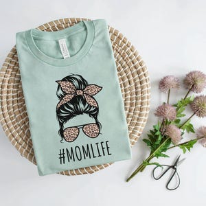 May include: A light green t-shirt with a graphic of a woman with a bun and sunglasses. The graphic has a leopard print design. The text "#MOMLIFE" is printed below the graphic.