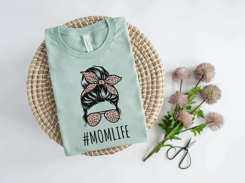 May include: A light green t-shirt with a graphic of a woman with a bun and sunglasses. The graphic has a leopard print design. The text "#MOMLIFE" is printed below the graphic.