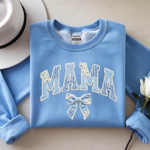 May include: A light blue sweatshirt with the word "MAMA" in a patchwork design, featuring floral, polka dot, and gingham patterns. A matching bow is below the word. A white hat and a white rose are also in the image.