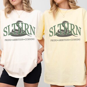 May include: Two t-shirts with a green snake graphic and the text "SLITHERN PRIDE AMBITION CUNNING". The shirt on the left is ivory and the shirt on the right is banana.