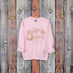 May include: Pink sweatshirt with a gold graphic of the Flourish and Blotts bookstore logo from the Harry Potter series. The logo says "Flourish and Blotts Booksellers Diagon Alley, London."