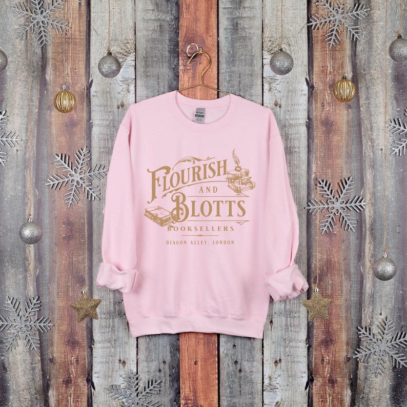 May include: Pink sweatshirt with a gold graphic of the Flourish and Blotts bookstore logo from the Harry Potter series. The logo says "Flourish and Blotts Booksellers Diagon Alley, London."