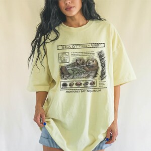 May include: Pale yellow t-shirt featuring a detailed graphic of sea otters with text that reads "Sea Otter - Enhydra Lutris" and "Monterey Bay Aquarium". The shirt has short sleeves and a relaxed fit.