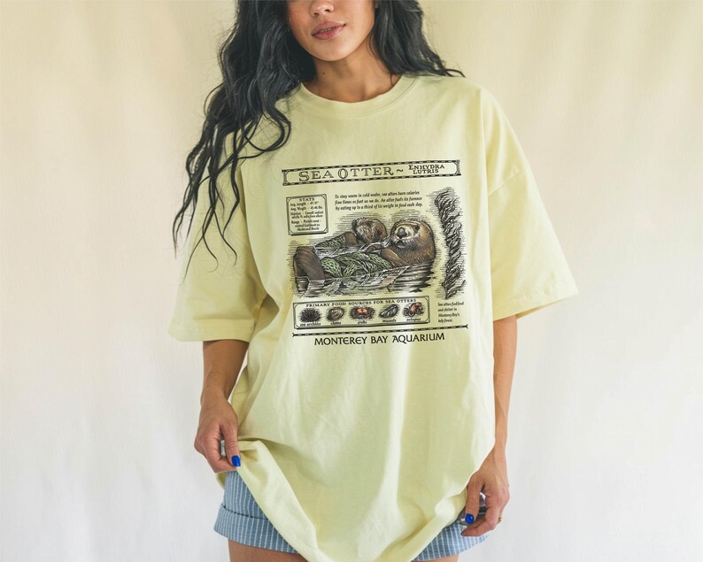 May include: Pale yellow t-shirt featuring a detailed graphic of sea otters with text that reads "Sea Otter - Enhydra Lutris" and "Monterey Bay Aquarium". The shirt has short sleeves and a relaxed fit.