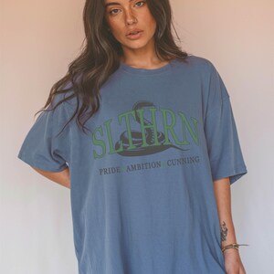 May include: A blue oversized t-shirt with a green snake graphic and the text "SLYTHERN" in green letters. Below the snake, the text "PRIDE AMBITION CUNNING" is printed in black.