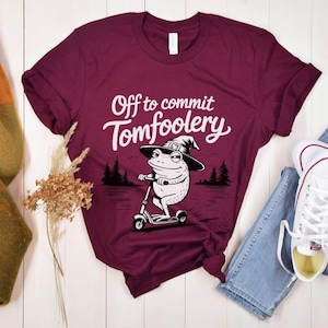 May include: A maroon t-shirt with the phrase "Off to commit Tomfoolery" in white script. The graphic features a frog wearing a witch hat and riding a scooter. The shirt is paired with blue jeans and white sneakers.