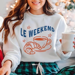 Może przedstawiać: Biała bluza z napisem "LE WEEKEND" w kolorze niebieskim, z pomarańczowymi ilustracjami filiżanki kawy, rogalika i gwiazd. Styl casual.