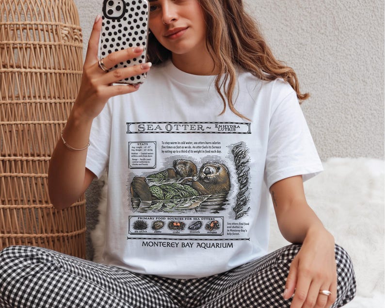 May include: White t-shirt featuring a detailed illustration of a sea otter with the text "SEA OTTER ~ Enhydra lutris". The shirt includes information about sea otters, their diet, and the Monterey Bay Aquarium.