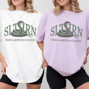 May include: Two t-shirts with a green snake graphic and the text "SLITHERN PRIDE AMBITION CUNNING". The shirt on the left is white and the shirt on the right is orchid.