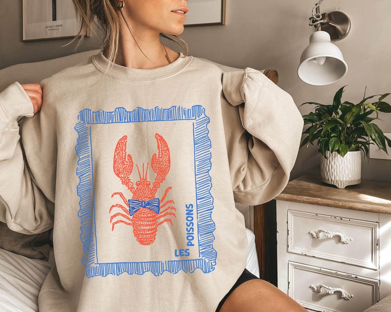 May include: Beige sweatshirt featuring a red lobster illustration with a blue bow tie and a blue frame. The text "LES POISSONS" is printed in blue. The sweatshirt has long sleeves and a crew neck.