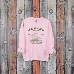 May include: Pink sweatshirt with a graphic of Ollivanders Wand Shop, established in 382 BC. The graphic features a book with a wand emerging from it, and the text "Makers of Fine Wands, Diagon Alley, London".