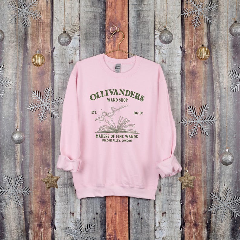 May include: Pink sweatshirt with a graphic of Ollivanders Wand Shop, established in 382 BC. The graphic features a book with a wand emerging from it, and the text "Makers of Fine Wands, Diagon Alley, London".