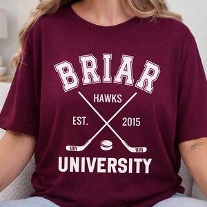 May include: Burgundy t-shirt with white text and graphics. The design includes the word "BRIAR" in a collegiate font, "HAWKS", crossed hockey sticks, a hockey puck, and "EST. 2015 UNIVERSITY".