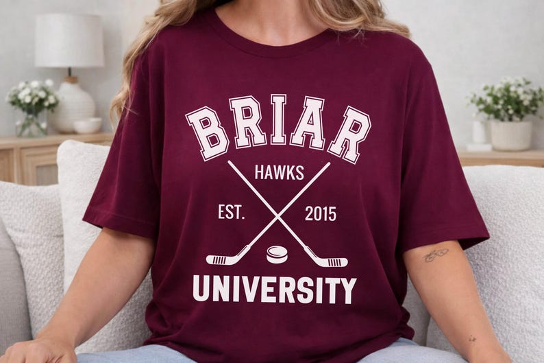May include: Burgundy t-shirt with white text and graphics. The design includes the word "BRIAR" in a collegiate font, "HAWKS", crossed hockey sticks, a hockey puck, and "EST. 2015 UNIVERSITY".