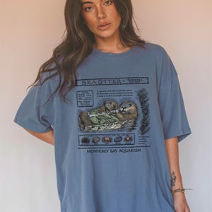 May include: A blue t-shirt featuring a detailed illustration of sea otters, with text that reads "SEA OTTER ~ ENHYDRA LUTRIS" and "Monterey Bay Aquarium." The graphic includes information about the otters and their diet.