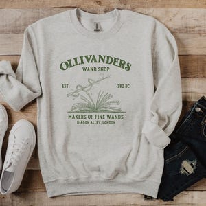 May include: A gray sweatshirt with a green graphic of Ollivanders Wand Shop. The graphic includes the text "Ollivanders Wand Shop", "Est. 382 BC", "Makers of Fine Wands", and "Diagon Alley, London".