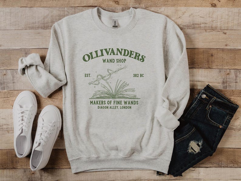 May include: A gray sweatshirt with a green graphic of Ollivanders Wand Shop. The graphic includes the text "Ollivanders Wand Shop", "Est. 382 BC", "Makers of Fine Wands", and "Diagon Alley, London".