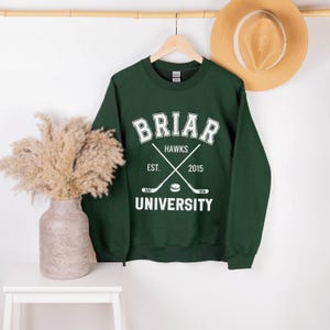 May include: A dark green sweatshirt with the text "Briar University Hawks" in white. The design includes crossed hockey sticks and a hockey puck. The sweatshirt is hanging on a wooden hanger.