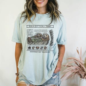 May include: Light blue t-shirt featuring a detailed illustration of sea otters with the text "Sea Otter - Enhydra Lutris" and "Monterey Bay Aquarium". The graphic includes scientific information and images of the otters' primary food sources.