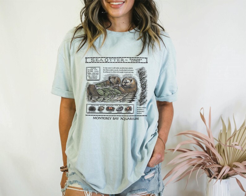 May include: Light blue t-shirt featuring a detailed illustration of sea otters with the text "Sea Otter - Enhydra Lutris" and "Monterey Bay Aquarium". The graphic includes scientific information and images of the otters' primary food sources.