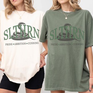 May include: Two t-shirts with a green snake graphic and the text "SLYTHERN PRIDE AMBITION CUNNING". The shirt on the left is ivory and the shirt on the right is moss.