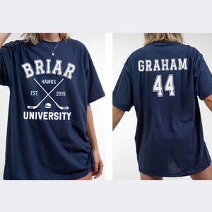 May include: Navy blue t-shirt with white text and graphics. The front features "Briar Hawks University" with crossed hockey sticks and the year "2015." The back displays "Graham 44." A casual, oversized fit.