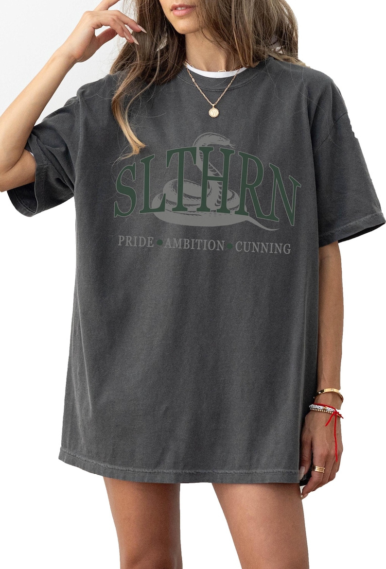 May include: A dark gray oversized t-shirt with a green snake graphic and the text "SLYTHERN" in white. Below the graphic is the text "PRIDE AMBITION CUNNING" in white.