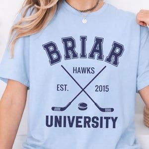 May include: Light blue t-shirt with a navy blue graphic. The graphic includes the word "BRIAR" above crossed hockey sticks and a puck, with "HAWKS" and "UNIVERSITY" below. The text "EST. 2015" is also included.