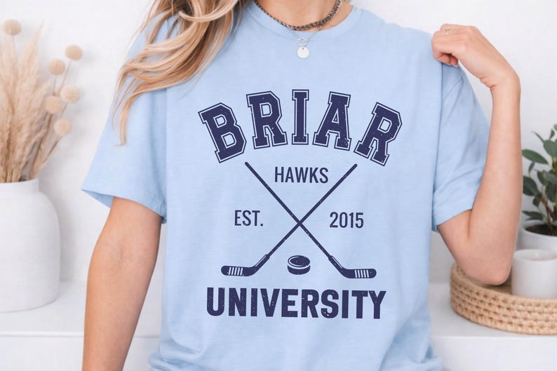 May include: Light blue t-shirt with a navy blue graphic. The graphic includes the word "BRIAR" above crossed hockey sticks and a puck, with "HAWKS" and "UNIVERSITY" below. The text "EST. 2015" is also included.