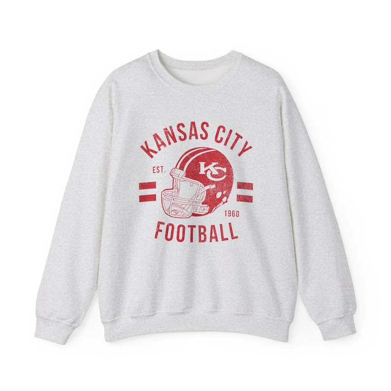 Pode incluir: Uma camisa de moletom cinza com um gr&aacute;fico vermelho de um capacete de futebol e o texto "Kansas City Football Est. 1960".
