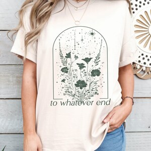 May include: A beige t-shirt with a graphic of a floral arrangement inside an arch with stars. The text "to whatever end" is printed below the graphic.