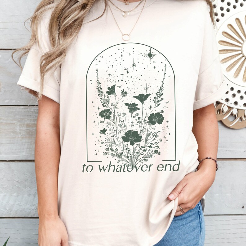 May include: A beige t-shirt with a graphic of a floral arrangement inside an arch with stars. The text "to whatever end" is printed below the graphic.