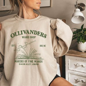 May include: A beige sweatshirt with a green graphic print of a wand, book, and the text "Ollivanders Wand Shop Est. 382 BC Makers of Fine Wands Diagon Alley, London"