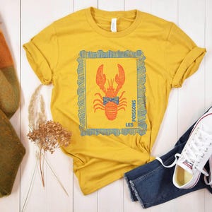 May include: Mustard yellow t-shirt with a lobster graphic in a blue frame. The lobster is orange with a blue bow tie. The text "LES POISSONS" is printed on the shirt. The shirt is paired with blue jeans and white sneakers.