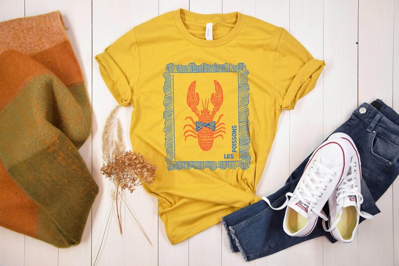 May include: Mustard yellow t-shirt with a lobster graphic in a blue frame. The lobster is orange with a blue bow tie. The text "LES POISSONS" is printed on the shirt. The shirt is paired with blue jeans and white sneakers.
